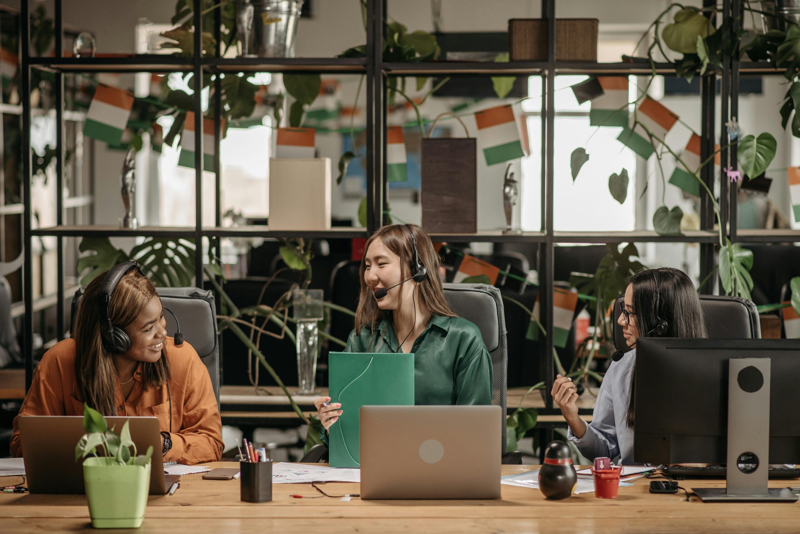 Three women with headsets working together in a vibrant, plant-filled modern office space.